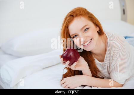 Glücklich rothaarige Frau auf dem Bett liegend und roten Apfel zu Hause halten Stockfoto