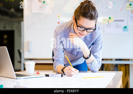 Hübsche junge Frau in Gläsern mit Laptop und Zeichnung Blaupause von Bleistift im Büro Stockfoto