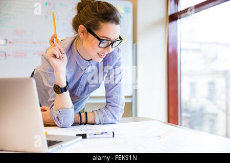 Glückliche junge Frau in Gläsern Stand in der Nähe der Fenster im Büro und arbeiten mit Bauplan Stockfoto