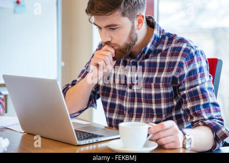 Serious konzentriert bärtigen jungen Mann im karierten Hemd am Tisch sitzen und mit laptop Stockfoto
