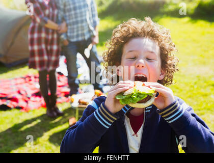 Begeisterte junge unter großen Bissen von Hamburger auf sonniger Campingplatz Stockfoto