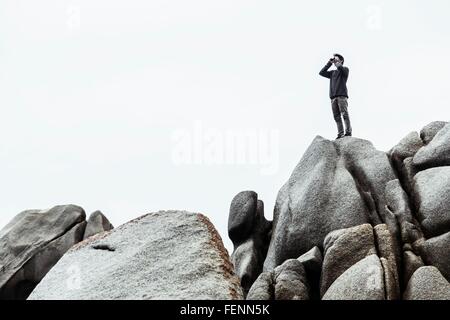 Niedrigen Winkel Blick des jungen Mannes stehen auf Felsen, vorgibt, verwenden Sie Fernglas, Costa Smeralda, Sardinien, Italien Stockfoto