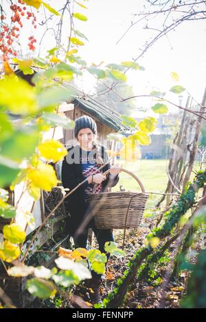 Blick durch die Blätter der jungen Frau im Garten tragen stricken Hut hält Korbwaren Korb Blick auf die Kamera zu Lächeln Stockfoto