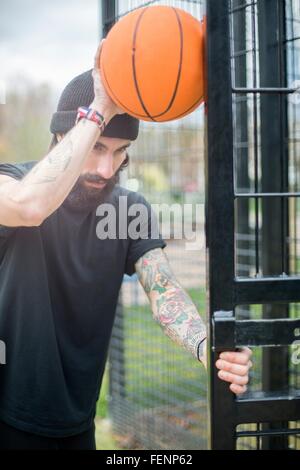 Mitte erwachsenen Mannes vom Zaun stehen, hält Basketball gegen Zaun, nachdenklichen Ausdruck Stockfoto