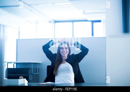 Junge Frau sitzt am Schreibtisch im Büro Hände hinter Kopf, Blick auf die Kamera zu Lächeln Stockfoto