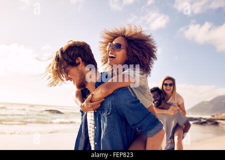 Gruppe von Freunden zu Fuß am Strand entlang, mit Männern geben Huckepack Fahrt an Freundinnen. Glückliche junge Freunde genießen einen Tag bei b Stockfoto