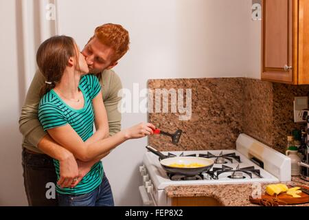 Junger Mann in der Küche Arme um junge Frau am Herd kochen, küssen Stockfoto