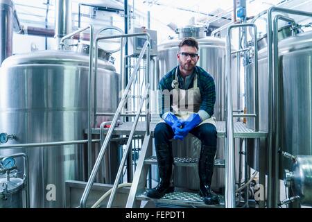 Junger Mann in der Brauerei tragen, Schürze, Handschuhe und Gummistiefel sitzen auf Stufen Blick in die Kamera Stockfoto