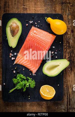 Frisches Lachsfilet, Zitronen, Avocados, Petersilie und Gewürzen auf schwarze Schneidebrett auf strukturierten hölzernen Hintergrund. Ansicht von oben Stockfoto
