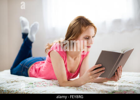 Entspannende Frau liest Buch mit einem Lächeln in weiß innen Stockfoto