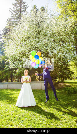 Bräutigam auf die Ballons fliegen, aber die Braut hält seine Hand. Lustige Hochzeit. Bräutigam springt und fliegt auf Ballons. Junger Mann von seiner Frau fliegen. Lustige Hochzeitskonzept. Happy Valentines day Stockfoto