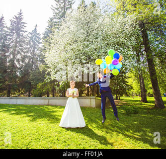 Bräutigam auf die Ballons fliegen, aber die Braut hält seine Hand. Lustige Hochzeit. Bräutigam springt und fliegt auf Ballons. Junger Mann von seiner Frau fliegen. Lustige Hochzeitskonzept. Happy Valentines day Stockfoto