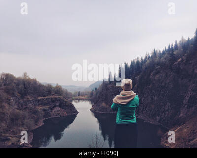 Ein Fotograf in grüner Jacke, Strickmütze und Schal fängt das Spiegelbild eines Teichs ein, der von Bäumen umgeben ist. Der bewölkte Himmel verleiht der Natur einen ruhigen und atmosphärischen Touch. Stockfoto