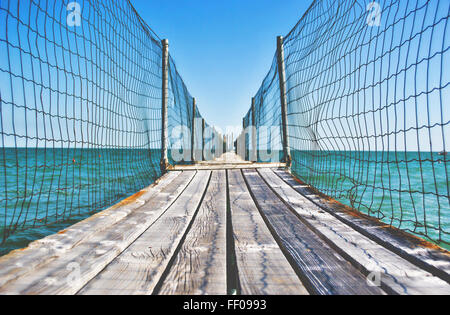 Eine umzäunte Holzpromenade erstreckt sich über das Meer, mit Blick auf blaues Wasser und klaren Himmel. Die Promenade bietet einen malerischen, ruhigen Pfad, auf dem Besucher die Küste aus einer erhöhten Perspektive genießen können. Der Zaun entlang der Promenade sorgt für Sicherheit, während Sie über das Meer laufen. Stockfoto
