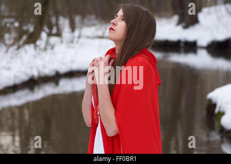 Ein Mädchen im roten Umhang steht nahe dem Fluss im winter Stockfoto