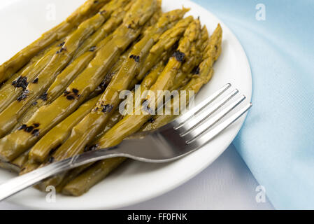 Eine Nahaufnahme von gegrilltem Spargel getränkt in Sonnenblumenöl auf einem weißen Teller plus Gabel auf einem off White Tischdecke und ein blau Stockfoto
