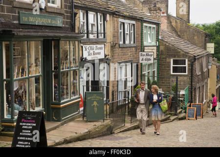 Paar zu Fuß bis steile Main Street, Haworth, West Yorkshire, England - malerischen, gepflasterten, gesäumt mit Geschäften, beliebt bei Bronte Land Besucher! Stockfoto