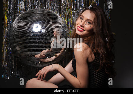 Nahaufnahme von schönen lächelnden jungen Frau mit funkelt in Haar und Gesicht posiert in der Nähe von Disco-Kugel über schwarzen Hintergrund Stockfoto