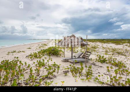 Gebrochene hölzerne Sonnenliegen und ein verlassener palm Leaf überdachte Sonnenschirm am Strand im Süden Barbuda, Antigua & Barbuda unter grauem Himmel Stockfoto