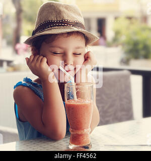 Spaß Kind Mädchen mit Hut Kniff ihre Augen und warten im Moment den Smoothie lecker Saft auf Sommer Hintergrund im Café zu trinken. Stockfoto