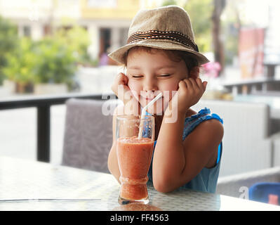 Spaß Kind Mädchen mit Hut Kniff ihre Augen und warten im Moment den Smoothie lecker Saft auf Sommer Hintergrund im Café zu trinken Stockfoto