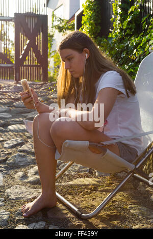 Junge Frau sitzt auf Liegestuhl Smartphone halten und hören Musik in einen Innenhof Hintergrundbeleuchtung mit Sonnenlicht in Schweden im Sommer Stockfoto