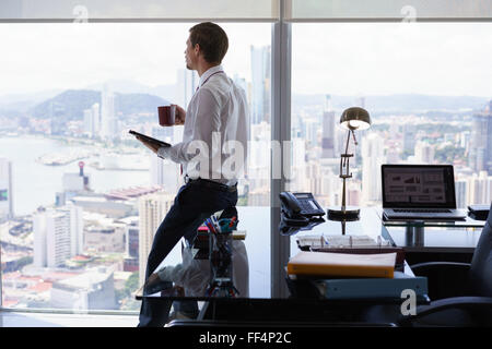 Erwachsenen Geschäftsmann auf Schreibtisch im modernen Büro sitzen und Lesen von Nachrichten auf Tablet-pc mit einer Tasse Kaffee. Der Mann sieht, der die Stockfoto