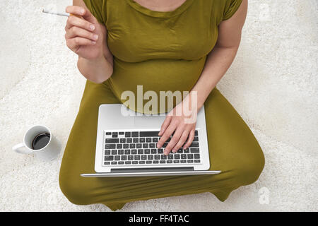 Junge schwangere Frau mit Zigarette und Laptop zu Hause entspannen Stockfoto