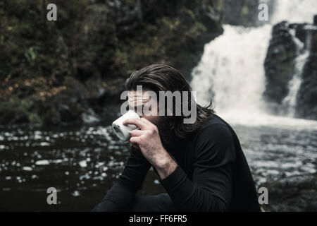 Wildes Campen. Ein Mann, trinken aus einer Tasse an einem schnell fließenden Bach. Stockfoto