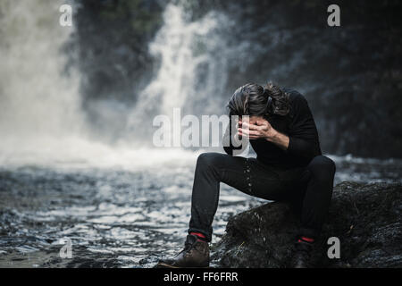 Ein Mann sitzt waschen Sie sein Gesicht in einem Gebirgsbach von einem Wasserfall. Stockfoto
