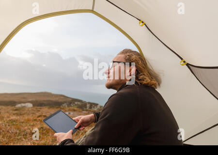 Ein Mann sitzt im Schutz eines Zeltes auf, hält eine digitale-Tablette. Stockfoto