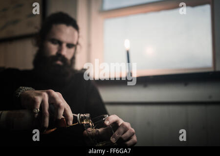 Ein Mann sitzt allein in einem Raum gießt sich ein Glas Whisky. Eine brennende Kerze. Stockfoto