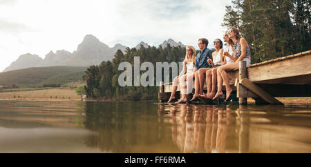 Porträt der glückliche junge Freunde sitzen auf Pier am See trinken Bier. Junge Männer und Frauen, die einen Tag am See zu genießen. Stockfoto