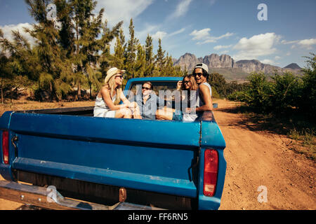 Gruppe von Freunden auf der Rückseite ein Pick up Auto sitzen. Junge Männer und Frauen, die auf einer Straße Reise in der Natur.