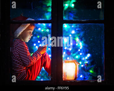 Netter Junge, sitzt auf einem Fenster Schild, spielen auf dem Handy in der Nacht, Weihnachten, Weihnachtsmann wartet Stockfoto