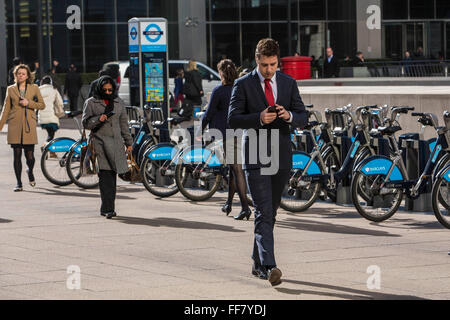 Ein Stadt-Arbeiter prüft seine Handy wie er über den Platz außerhalb Canary Wharf u-Bahnstation in den Docklands, London, Vereinigtes Königreich geht. Er geht vorbei an Barclays-Bike-Park, den Spitznamen Boris Bikes, die sind eine beliebte Form des Transports für Londoner. Stockfoto