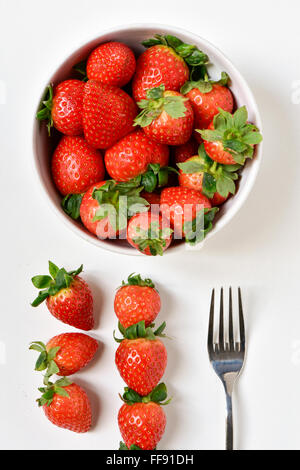 High-Angle Shot einer weißen Keramik Schüssel voll appetitlich Erdbeeren, eine Gabel und einige andere Erdbeeren auf einer weißen Fläche Stockfoto