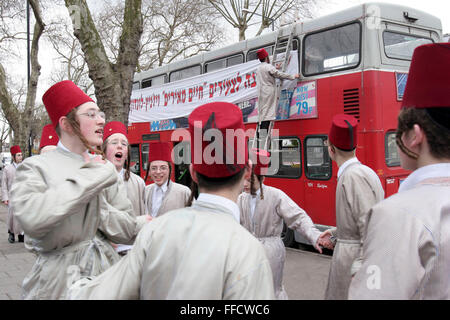 Eine Gruppe von orthodoxen jüdischen Jungen aus der Viznitz-Jeschiwa (Schule) in Kostüm gekleidet singen und tanzen auf der Straße. Andere Mitglieder der Gruppe schmücken den Doppeldecker-Bus mit Bannern, die sie für die jüdischen Purim-Festival auf Reisen werden. Sie werden mehrere lokale Wohlhabende Geschäftsleute sammeln Geld für ihre Schule besuchen. Stockfoto