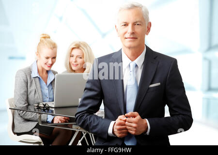 Porträt von senior Geschäftsmann im Büro während der Unternehmerinnen stehen im Hintergrund vor Laptop stehen. Stockfoto