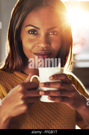 Attraktive schwarze Frau, die bei einem energiespendenden Tasse Kaffee hält den Becher in der Hand als sie schaut die Kamera mit einem leisen Stockfoto