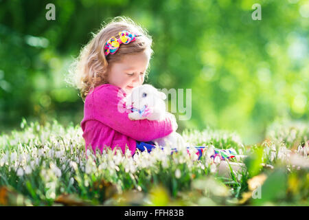Niedlichen Kleinkind Mädchen spielen mit echten Hasen in blühenden Frühling Garten. Kind auf Ostereiersuche. Kind mit bunten Eiern und streicheln Stockfoto