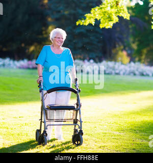 Glücklich senior Behinderte Dame mit einer Gehbehinderung, genießen Sie einen Spaziergang in einem sonnigen Park schieben ihre Gehhilfe oder Rollstuhl Stockfoto