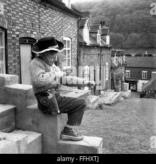 Kleiner Junge, der alleine mit einem Katapult spielt und ein Cowboy-Outfit trägt 1960s Britain UK Street vor den Straßen Stockfoto