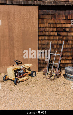 Farm Equipment Stilleben vor ein landwirtschaftliches Gebäude mit einem kleinen vierrädrigen Traktor, antike zwei Rädern Sackkarre platziert. Stockfoto