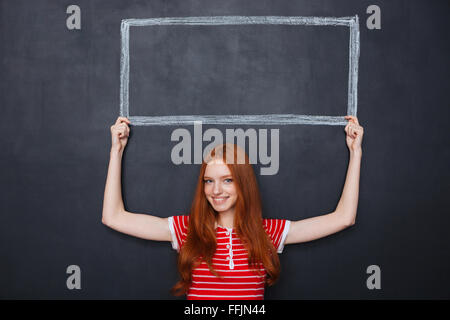 Attraktive lächelnd rothaarige junge Frau Halterahmen auf Tafel Hintergrund über den Kopf gezogen Stockfoto