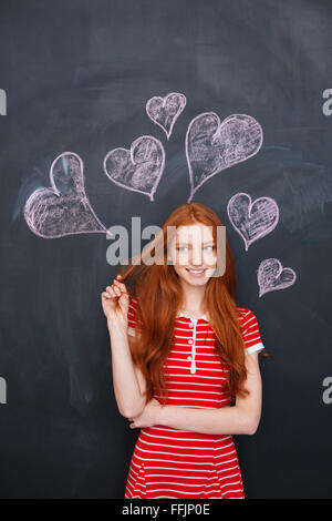 Attraktive rothaarige junge Frau über Blackboard mit gezeichneten Herzen hinter ihr stehen Stockfoto