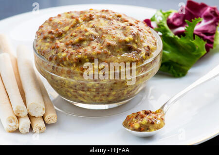 Dijon-Senf mit Brot-Sticks und Salat. Fokus auf vorderen Senf in einem kleinen silbernen Löffel auf einem weißen Porzellanteller Stockfoto