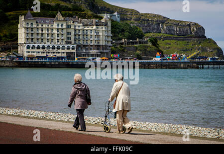 Zwei ältere Damen, die zu Fuß entlang der Promenade in Llandudno mit The Grand Hotel und The Great Orme im Hintergrund Stockfoto