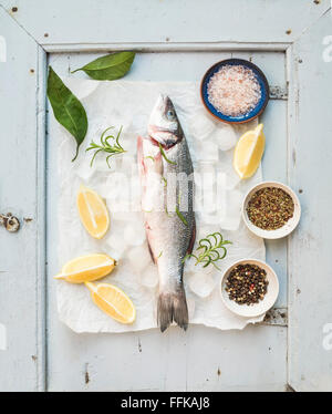 Frische ungekochte mediterranen Wolfsbarsch Fisch mit Zitrone, Kräutern, Eis und Gewürzen auf rustikalen blau Holzbrett Kulisse, Ansicht von oben Stockfoto