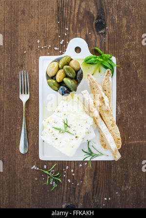 Frischer Schafskäse mit Oliven, Basilikum, Rosmarin und Brot Scheiben auf weißer Keramik dient Board über rustikalen hölzernen Hintergrund Stockfoto